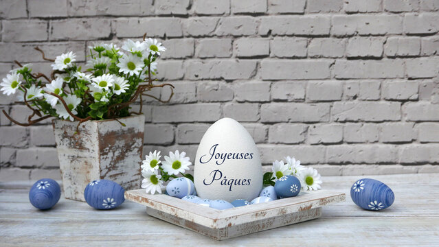 Carte de v&oelig;ux pour P&acirc;ques&nbsp;: Nid de P&acirc;ques avec des &oelig;ufs bleus et des fleurs devant un mur de pierre. Un &oelig;uf porte l&rsquo;inscription &laquo;&nbsp;Joyeuses P&acirc;ques&nbsp;&raquo;.