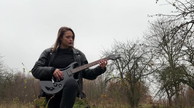 A female rock musician in a leather jacket plays an electric guitar with an amplifier in the garden in gloomy weather