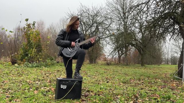 young female rock musician in a leather jacket plays an electric guitar outdoors