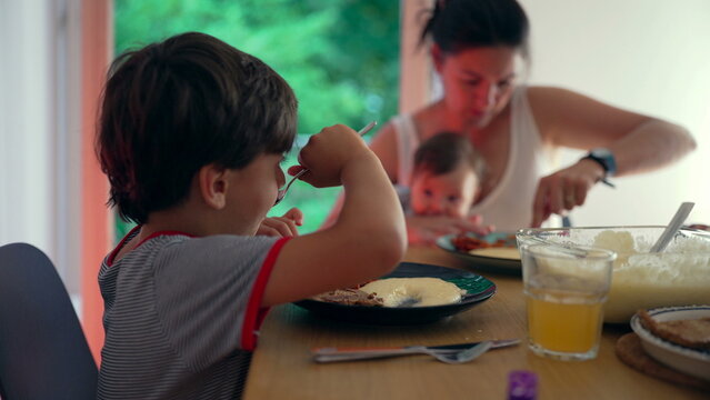 Family dining together, mother multitasking while older child eats independently, home-cooked meal, nurturing environment, parent balancing caregiving and mealtime