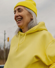 Elderly woman laughing outdoors in yellow beanie and hoodie