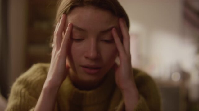 Handheld close up shot of distressed young adult woman holding head with hands experiencing intense anxiety while sitting in bedroom, subtle camera shake effect