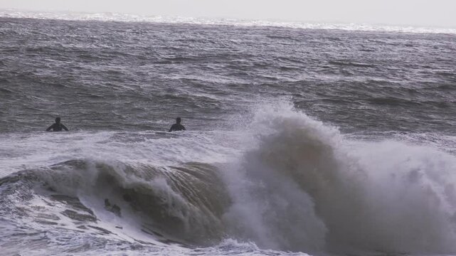 Wide shot of surfers waiting beyond breaking waves in the Atlantic Ocean. Choppy water and sea spray create a dramatic coastal mood.