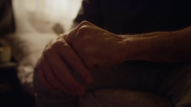 Close up on arms of unrecognizable middle aged man rubbing hands soothing tremor in fingers struggling with stress in low key lit bedroom