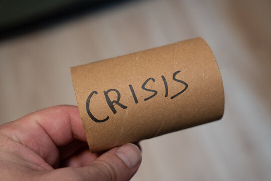 A hand holds a brown cardboard cylinder with the word CRISIS handwritten on it, symbolizing emergency, difficulty, or a challenging situation. This image represents economic, social, or personal chal