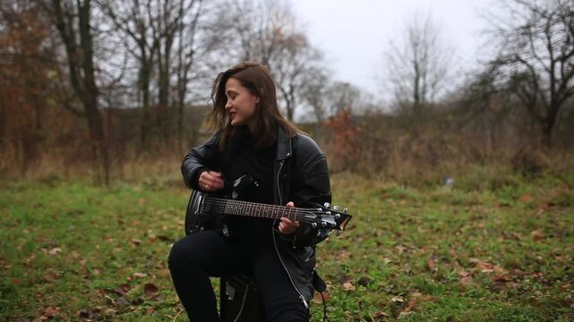 A female rock musician in a leather jacket plays an electric guitar with an amplifier in the garden in gloomy weather
