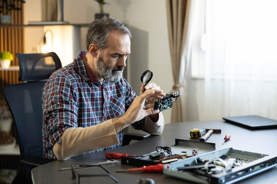 Man examining a circuit board with a magnifying glass while repairing consumer electronics at a home workshop