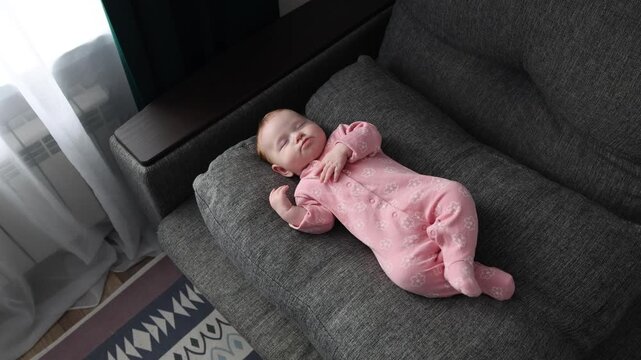 Infant in pink floral pajamas wakes up on a grey sofa, stretching arms and rubbing eyes in the morning light