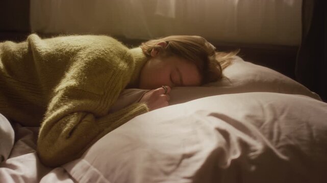 Tilt up shot of overwhelmed young adult woman lying down on bed pillow with eyes closed experiencing emotional crisis in bedroom filled with sunlight