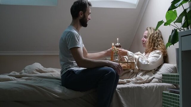 Young married couple celebrating an anniversary in bed. The husband brings breakfast in bed, sits beside his wife and gives her a cupcake with a candle