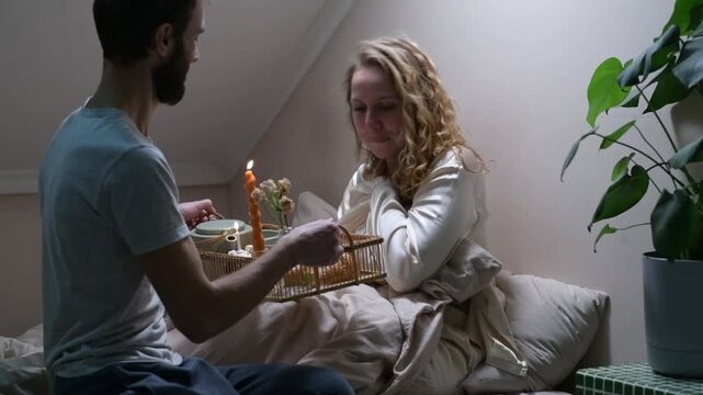 Young married couple celebrating an anniversary in bed. The husband brings breakfast in bed, sits beside his wife and gives her a cupcake with a candle