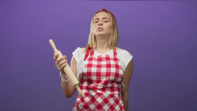 Woman chef in red gingham apron holding a rolling pin and flicks her wrist while looking upward in studio; impatience.