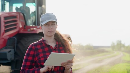 Woman farm owner managers farm using tablet. Woman farmer works with tablet in field against background of tractor. Agriculture. Worker on tractor cultivates and fertilizes field. Tractor works field © Victoriia