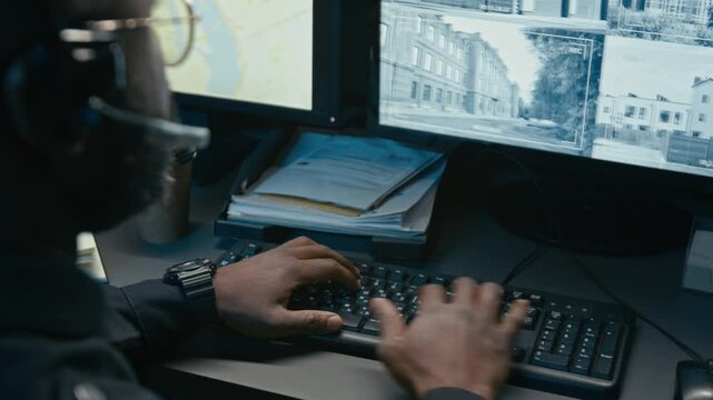Over shoulder close up shot of unrecognizable Black male police officer and using computer at desk with focus on hands typing on keyboard during night shift