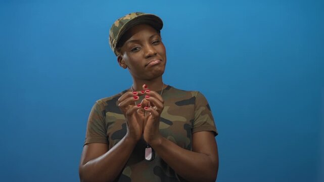 Young african american woman in camouflage shirt claps hands while wearing dog tag necklace in blue studio; pride duty honor discipline.