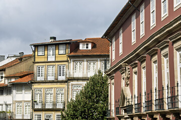 Fototapeta premium A red and yellow facade in Guimarães rises with white frame window and ornate trim, the tree and soft sky giving the street a calm historic charm.