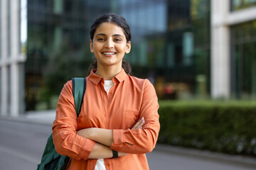 Young indian woman student standing outdoors with a backpack and crossed arms, confidently smiling...