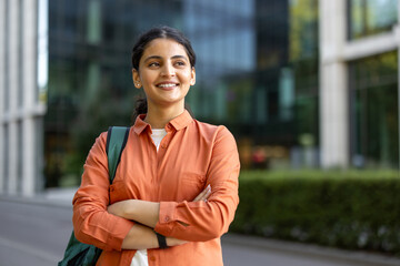 Naklejka premium Young indian woman carrying a backpack and crossing arms, looking away and smiling in front of a modern university building, representing education, success, and future opportunities
