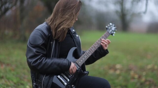 A female rock musician in a leather jacket plays an electric guitar outdoors in gloomy weather