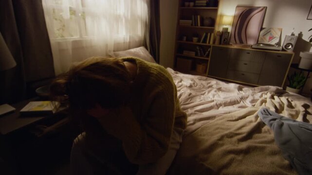 Desperate young adult woman sitting on bedside holding head while experiencing intense anxiety then falling onto bed in sunlit room
