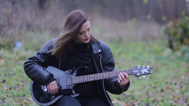 A female rock musician in a leather jacket plays an electric guitar outdoors in gloomy weather