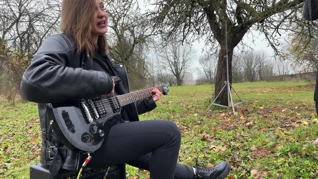 A woman plays an electric guitar outdoors while sitting on a speaker in gloomy weather.