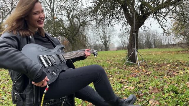 A woman plays an electric guitar outdoors while sitting on a speaker in gloomy weather.