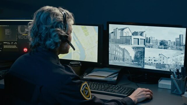 Back view shot of grey haired woman working as dispatcher at police command center during night shift and speaking to headset while watching computer monitors with city street views, copy space