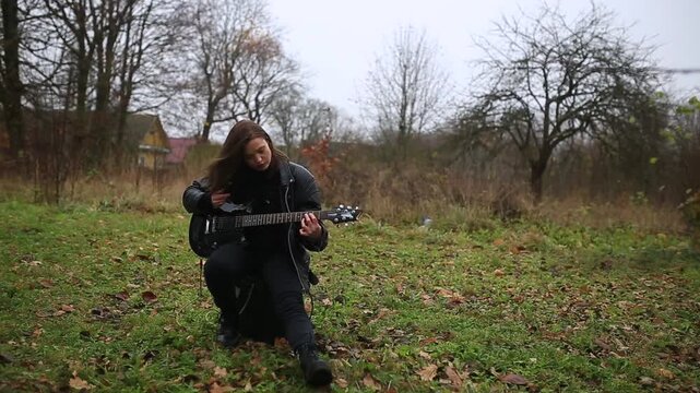 A female rock musician in a leather jacket plays an electric guitar with an amplifier in the garden in gloomy weather
