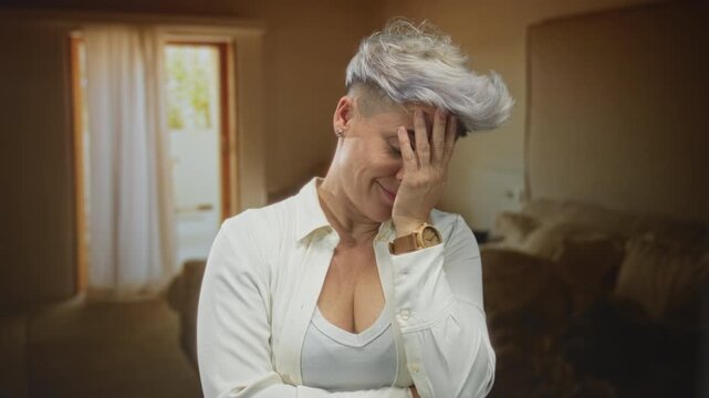 Woman with short hair wearing white shirt holds hand to forehead in building, standing by doorway and curtain; embarrassment.