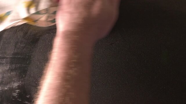 A man's hand, holding a rag, cleaning the dust off a flat, plain, black surface, by moving the hand back and forth. 