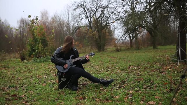 A woman plays an electric guitar outdoors while sitting on a speaker in gloomy weather.