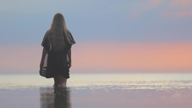 A girl with blond chic hair in a black bikini and a lace shawl walks along the estuary