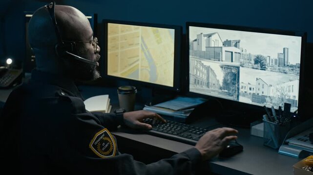 Side view shot of African American man working as dispatcher at police station and using computer while reviewing CCTV feed of city streets on monitors during night shift