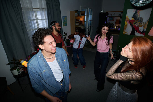 Group of young people and teenagers, including woman, Black man, and multiethnic friends, laughing and dancing together in living room during lively house party gathering