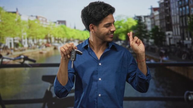 Man holding car keys with fingers crossed on street, smiling in a casual blue shirt near canal railing, bare hands visible; hope fresh start.