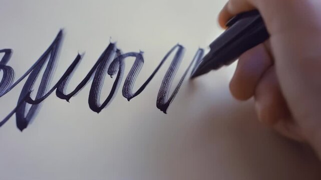 Hand of a young adult female artist using a black marker to create elegant calligraphy strokes on a smooth sheet of paper in a well-lit indoor setting