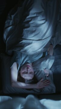 Vertical top view shot of young adult woman rolling from side to side on bed attempting to sleep then tossing pillow across bed nervously during night