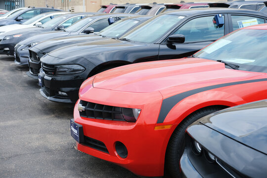 Used cars parked at a dealership lot showing a red sports car front and multiple black and gray sedans lined up ready for sale, and customer test drive