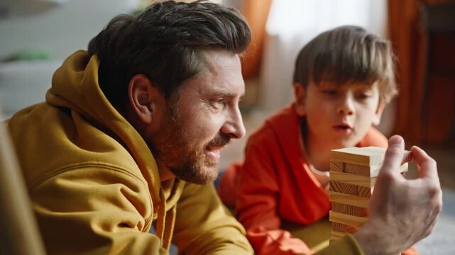 Father taking jenga block playing lovely little son lying room floor closeup.
