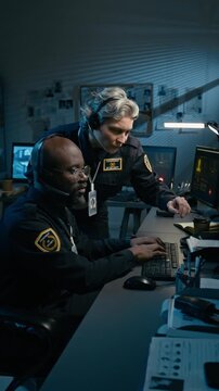 Vertical side view shot of two police officers discussing operation in control room while working night shift and pointing at screen using computer together