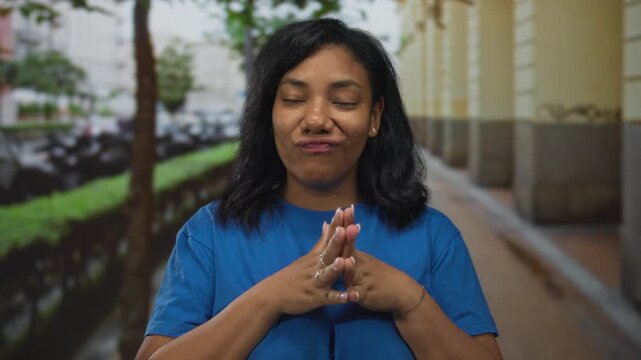 Woman wearing blue shirt steeples fingers in confidence gesture on tree lined city street; confidence assurance certainty poise.