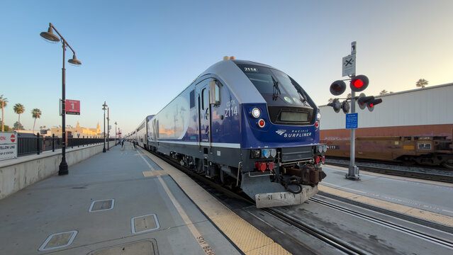 LOS ANGELES, CA, NOV 8, 2025: Amtrak Pacific Surfliner train sits at a station platform at dusk, with red crossing signals illuminated and palm trees in background