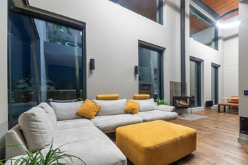 Fototapeta premium Living room in a country house decorated in high-tech style. Modern architectural design, high ceiling, panoramic windows with a view of the evening winter landscape.