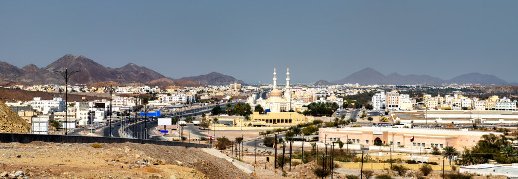A panoramic cityscape of Rustaq, Oman, showcasing the Sultan Qaboos Mosque and the rugged Hajar Mountains under a clear blue sky.