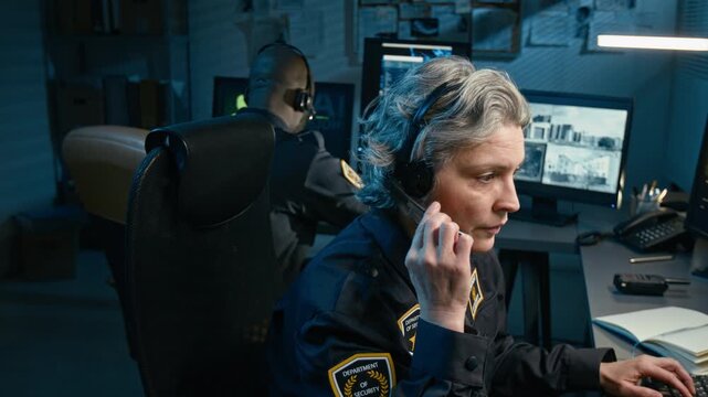 Cinematic side view shot of adult female dispatcher at police station and answering calls via headset while sitting at desk and watching monitors at night