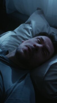 Vertical shot of adult man lying awake at night pressing hands to face in annoyance then turning over onto pillow trying to get comfortable and sleep during late night