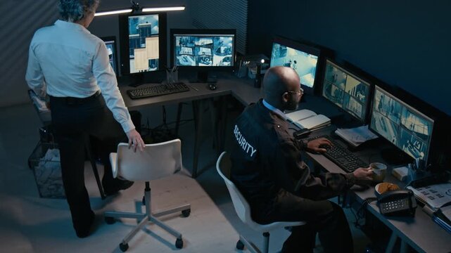 High angle shot of two security officers sitting at shared workstation and using multiple computer monitors showing CCTV footage during night shift
