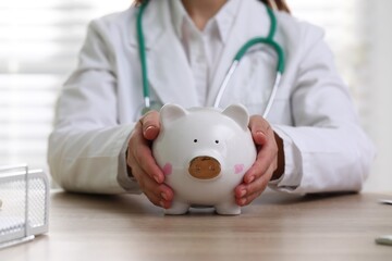 Doctor in medical coat with white piggy bank at wooden table indoors, closeup