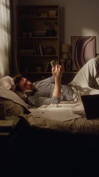 Vertical shot of adult man lying on bed in dimly lit room scrolling phone, struggling to focus on paperwork while avoiding work tasks